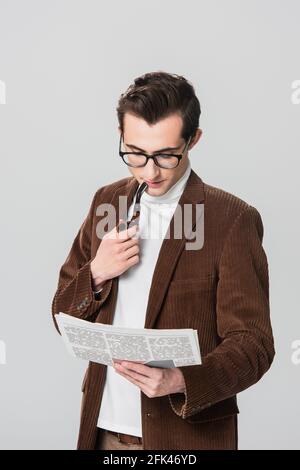 Junger Mann in Samtblazer raucht Pfeife, während er Zeitung liest Isoliert auf Grau Stockfoto