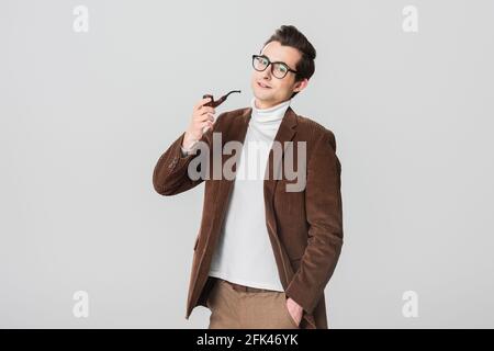 Stilvoller Mann in altmodischem Blazer raucht Pfeife beim Posen Mit Hand in Tasche isoliert auf grau Stockfoto