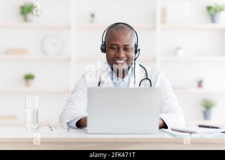 Online-Konsultation während der Quarantäne. Fröhlicher schwarzer Arzt, der Kopfhörer trägt und über einen Laptop mit dem Patienten spricht Stockfoto