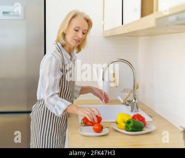 Glückliche ältere Frau Großmutter in Schürze waschen frisches Gemüse für Salat, während genießen Kochen in modernen hellen Küche zu Hause. Lächelnde ältere Frau, die gesunde Lebensmittel für die Familie vorbereitet Stockfoto