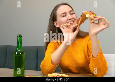 Die hübsche junge dunkelhaarige Frau in einem orangefarbenen Sweatshirt hält mit beiden Händen und einem Lächeln ein Stück Pizza neben einem Bier in einer grünen Flasche Stockfoto