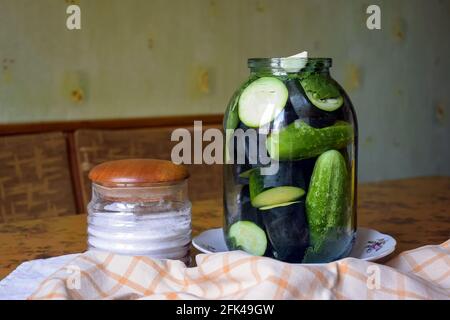 Hausgemachtes Beizen. Auf dem Küchentisch Glas mit frischen Gurken und eingelegten Zutaten. In der Nähe befindet sich ein Glasbehälter mit Salz. Nahaufnahme. Stockfoto