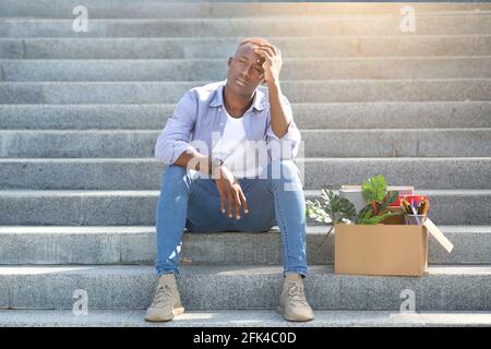 Krise und Arbeitslosigkeit. Gestresster schwarzer Kerl mit persönlichen Sachen, die auf Treppen sitzen, nachdem er seinen Job verloren hat, im Freien Stockfoto