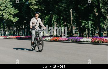 Fröhlicher junger Mitarbeiter mit Helm auf dem Fahrrad fährt zu Moderner Arbeitsplatz Stockfoto