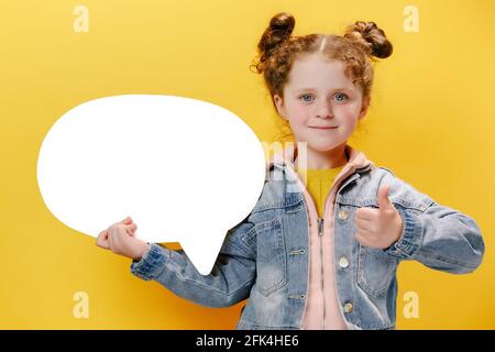 Nahaufnahme Porträt eines glücklichen kleinen Mädchens, das große weiße Sprechblase mit Daumen nach oben hält, wie Geste, gekleidet in eine blaue Jeansjacke Stockfoto