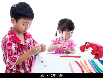 Kleine asiatische Kinder spielen und erstellen Spielzeug aus Spielteig auf dem Tisch. Bezauberndes Mädchen, das auf roten Dinosaurier-Ton-Modell, auf weißem Hintergrund. Stren Stockfoto