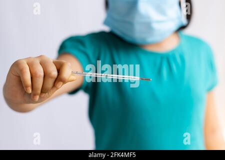 Selektiver Fokus auf ein analoges Thermometer, das in der Hand eines Kindes gehalten wird, das eine chirurgische Nasenmaske trägt, Gesundheitskonzept Stockfoto