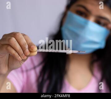Selektiver Fokus auf ein analoges Thermometer, das in der Hand einer mittleren erwachsenen indischen Frau gehalten wird, die eine chirurgische Nasenmaske trägt und die Stirn berührt, Gesundheitsvorsorge concep Stockfoto