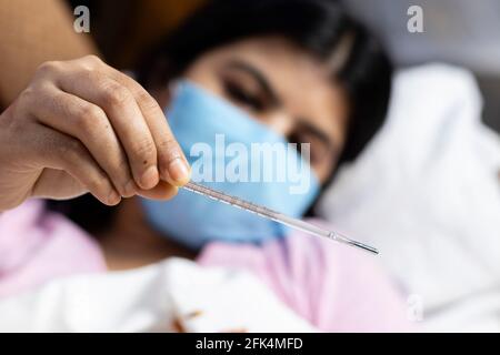 Selektiver Fokus auf ein nahes analoges Thermometer, das in der Hand einer mittleren erwachsenen indischen Frau gehalten wird, die eine chirurgische Nasenmaske trägt und auf dem Sofa liegt, Gesundheitsfürsorge-CO Stockfoto