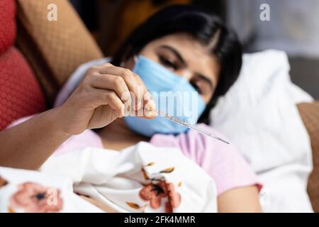 Selektiver Fokus auf ein analoges Thermometer, das in der Hand einer mittleren erwachsenen indischen Frau gehalten wird, die eine chirurgische Nasenmaske trägt und auf einem Sofa liegt, Gesundheitskonzept Stockfoto