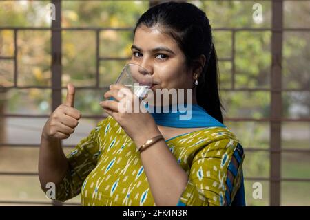 Eine indisch-asiatische Frau, die Wasser aus Glas trinkt und zeigt Daumen hoch Stockfoto