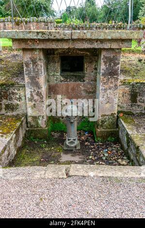 Ein alter Trinkwasserbrunnen aus Gusseisen, zylindrisch mit Längsrippen, eine Kuppel oben und ein Löwenkopf-Auslauf unter einem Steinhaus Stockfoto
