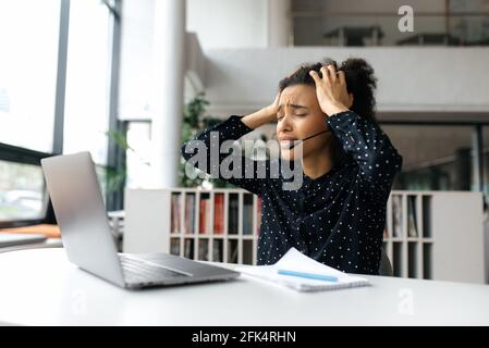 Verärgert müde afroamerikanische junge lockige Frau, mit Headset, Manager, Callcenter-Mitarbeiter oder Support-Operator, sitzt am Schreibtisch, fühlt sich gestresst, hält den Kopf mit den Händen und schreit Stockfoto