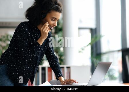 Intelligente, selbstbewusste, junge afroamerikanische Frau im Headset, Manager, Callcenter-Mitarbeiter oder Support-Mitarbeiter, die neben dem Schreibtisch steht, mit dem Kunden spricht, mit dem Kunden konsultiert und lächelt Stockfoto