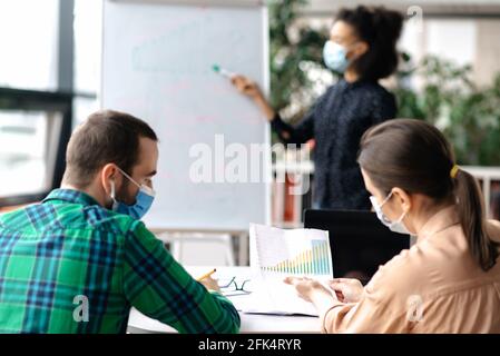 Erfolgreiche Mitarbeiter versammelten sich im Büro in schützenden medizinischen Masken, diskutieren ein Business-Projekt, vor dem Hintergrund verschwommen afroamerikanische Frau zeigt Informationen auf Whiteboard Stockfoto