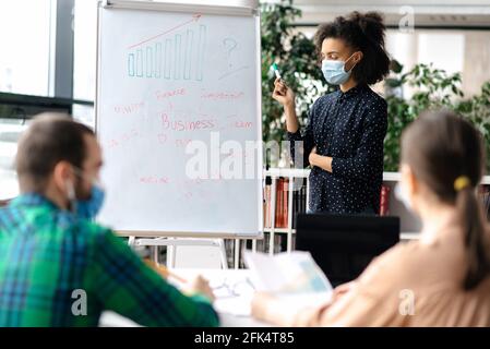 Business smart junge afroamerikanische Frau, trägt schützende medizinische Maske steht in der Nähe Whiteboard im Büro, stilvoll gekleidet, zeigt und erklärt Business-Plan und Strategie zu ihren Kollegen Stockfoto