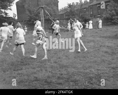 Die 50er Jahre, historisch, auf einem Stück Land am Ende der Terrasse viktorianischer Hütten, eine Gruppe von lokalen Männern - "The Heavy Gang" in Frauen gekleidet tanzen um eine Maypole, England, Großbritannien. Der Maifeiertag war eine uralte Tradition, die die Ankunft von Frühling und Sommer feierte, und Darsteller reenact alte Folklore und Musik, mit dem Springen um einen hohen hölzernen Pol, ein Maibaum, eine der populären Feier Aktivitäten. Stockfoto