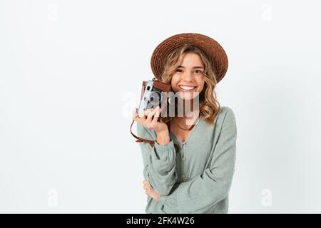 Glückliche junge Fotografin mit Hut hält ihre Kamera lächelnd auf weißem Studiohintergrund. Stockfoto