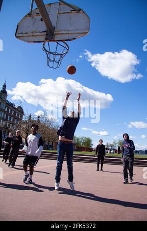 Multiethnische junge Menschen zahlen Straßenkbasketball. Der Ball liegt in der Luft und die Spieler werden ausgespielt. Blauer Himmel und Gebäude im Hintergrund. Kopenhagen Stockfoto