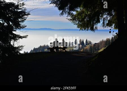 Pärchen sitzt auf einer Bank in Silhouette, eingerahmt von Bäumen, und beobachtet den Sonnenuntergang in den Bergen Stockfoto