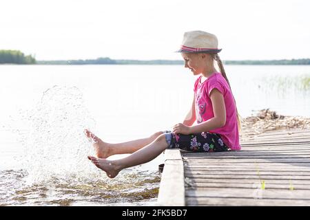 Ein glückliches kleines Mädchen in einem Strohhut sitzt auf einem hölzernen Ponton auf dem See. Stockfoto