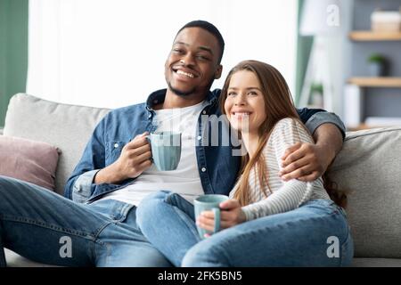 Inlandswochenende. Glückliches multikulturelles Paar, das zu Hause Fernsehen und Kaffee trinken sieht Stockfoto