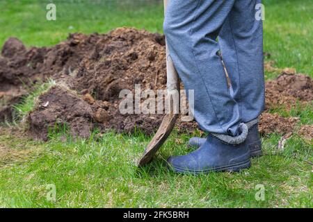 Beine eines Mannes in Gummistiefeln mit einer Schaufel in der Nähe des ausgegrabenen Bodens aus der Nähe. Stockfoto