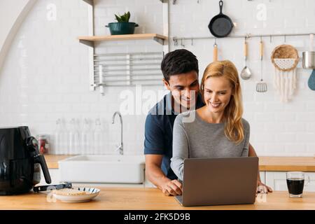 Romantische junge kaukasische Ehemann und Ehefrau mit Laptop während der Vorbereitung Gesunde Snacks in der Airfryer und Tee zu Hause trinken Stockfoto