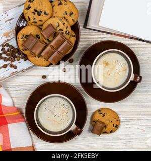 Zwei Tassen Kaffee mit Schokoladenkeksen und Schokolade auf dem Hintergrund eines offenen Buches. Heißer Kaffee mit Schokoladenkeksen im Tee Stockfoto