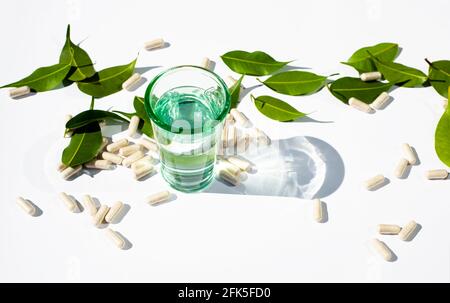 Glasbecher mit Wasser und Kapseln und Pillen auf weißem Hintergrund. Grüne Blätter. . Alternative Medizin Kopieren Sie Leerzeichen für Text. Stockfoto