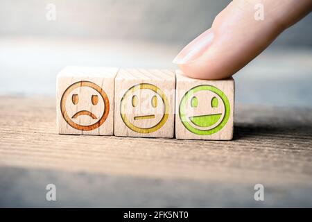 Gutes Feedback-Konzept mit roten, gelben und grünen Smileys auf Holzblöcken auf EINEM Brett wird der grüne Smiley von EINEM Finger berührt Stockfoto