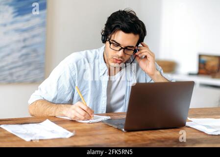 Konzentrierter, kluger junger hispanischer Mann mit Brille, Manager, Student oder Freiberufler, der zu Hause auf einem Laptop arbeitet, mit Mitarbeitern oder Kunden über ein Headset kommuniziert, während er Notizen macht Stockfoto