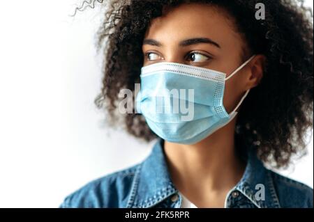 Quarantänevorkehrungen, Gesundheitskonzept. Nahaufnahme eines Porträts einer jungen afroamerikanischen, lockigen Brünette, die drinnen steht, mit einer schützenden medizinischen Maske auf ihrem Gesicht, die zur Seite schaut Stockfoto
