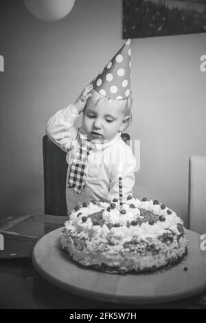 Vertikale Graustufenaufnahme eines kaukasischen Kleinkindes mit einem Geburtstagskappe in der Nähe seines Geburtstagstorte Stockfoto