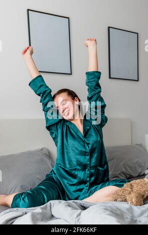 Junge Frau im Seiden-Pyjama gähnt, während sie in einem bequemen weichen Bett sitzt. Stockfoto