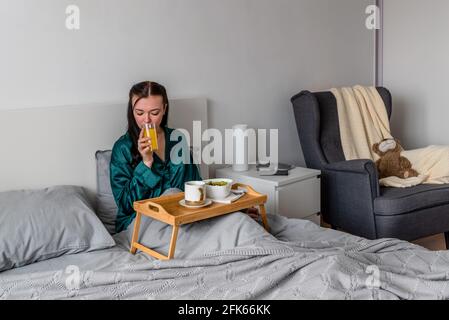 Junge Frau, die im Bett liegt, isst ihr Frühstück und trinkt frisch gepressten Saft. Wellness-Konzept. Stockfoto