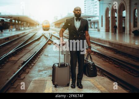 Ein ernsthafter bärtiger, haarloser, gut gekleideter Schwarzmann-Unternehmer in einem Anzug mit Fliege und Gepäck wartet auf dem Die Plattform des Bahnhofs d Stockfoto