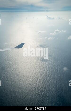 Vertikale Aufnahme aus dem Flugzeugfenster mit dem Ozean, der von seltenen winzigen Wolken bedeckt ist und einem Flugzeugflügel, der während des Drehens nach unten geneigt ist; nebliger Horizont, Wellen Stockfoto