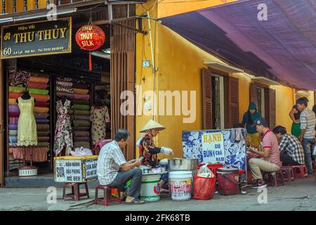 Vietnamesische Frau, die Street Food im Straßencafé neben dem Stoffladen in der Altstadt, Hoi an, Vietnam, verkauft Stockfoto