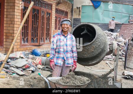 Fröhliche vietnamesische Handarbeiterin, die Beton im Zementmischer auf der Baustelle, Hoi an, Vietnam, mischt Stockfoto