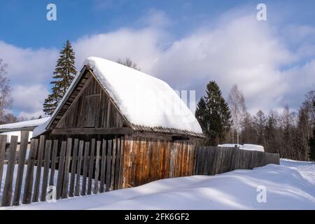 Ein Holzschuppen mit weißem Schnee bedeckt und ein langer Zaun aus Planken an einem sonnigen Wintertag, Russland. Stockfoto