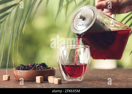 Frau gießt leckeren Hibiskustee aus der Teekane in Glas Tisch im Freien Stockfoto