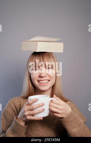 Nerd, Wissens- oder Bildungskonzept. Glückliche attraktive kaukasische Universitätsstudentin mit Brillen und beigem Pullover mit Buch auf dem Kopf und einer Tasse Kaffee oder Tee in den Händen auf grauem Hintergrund Stockfoto