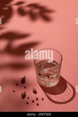 Kristallglas mit Gin und Tonic, garniert mit Rosenknospen und roten Pfefferkörnern auf pastellrosa Hintergrund. Schatten über die Szene setzen. Minimaler Sommer Stockfoto