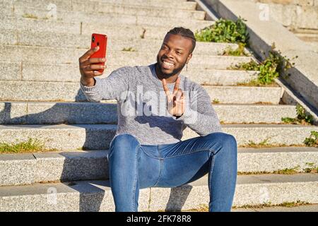 Junger afroamerikanischer Mann, der auf der Treppe sitzt und mit einem Smartphone ein Selbstporträt abnimmt. Glücklicher schwarzer Mann. Neue Normalität. Hochwertige Fotos Stockfoto