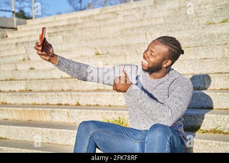 Junger afroamerikanischer Mann, der auf der Treppe sitzt und mit einem Smartphone ein Selbstporträt abnimmt. Glücklicher schwarzer Mann. Neue Normalität. Hochwertige Fotos Stockfoto