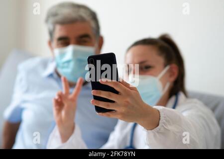Ärztin mit Schutzmaske feiert die Genesung des Patienten durch ein Selfie zusammen. Konzept des Erfolgs während der häuslichen Pflege. Konzentrieren Sie sich auf s Stockfoto
