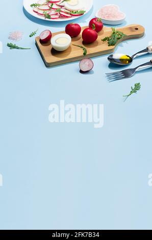 Diätsalat aus frischem Gemüse - Rettich, Rucola und Eier mit Schatten in hartem Licht auf blauem Hintergrund und weißem Holztisch, Rand, vertikal. Stockfoto