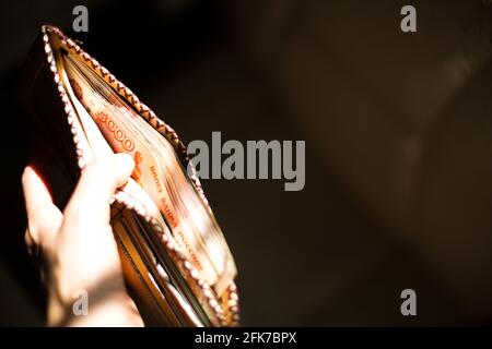 Leder-Brieftasche voller russischem Geld-5000 Scheine, fünftausend Rubel. Die Hand eines Mannes hält eine offene, überfließende Brieftasche vor einem schwarzen Hintergrund in einem Stockfoto