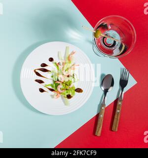 Frühlings-Seafood-Salat aus Garnelen, Grüns, Sellerie, Balsamico-Sauce mit martini-Cocktail in weißer Platte auf rotem und minzfarbenem Hintergrund mit Schatten Stockfoto